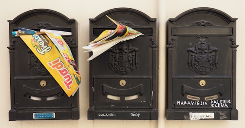 Three black mailboxes are mounted on a wall. Each mailbox displays a decorative emblem. Printed materials and advertisements are stuffed in the gaps of two mailboxes. Names are written on the bottom of each mailbox.