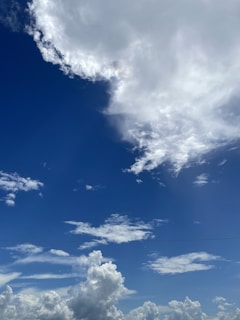 A clear sky dominated by large, fluffy clouds. The clouds are mostly white with some areas appearing more dense and gray. The background is a deep blue sky, creating a vivid contrast with the white cloud formations.