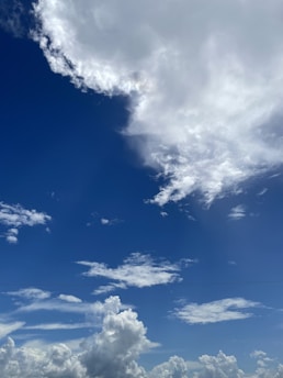 A clear sky dominated by large, fluffy clouds. The clouds are mostly white with some areas appearing more dense and gray. The background is a deep blue sky, creating a vivid contrast with the white cloud formations.
