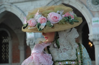 A person dressed in an elaborate, historical costume with a large, decorative hat adorned with pink roses and green leaves. The outfit includes intricate lace and satin fabrics in pastel shades, and the background features architectural elements with arches and brickwork.