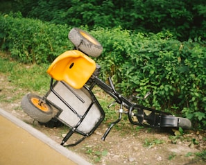 A small orange and black pedal-powered go-kart is overturned on a grassy area beside a paved path. It rests on its side with its wheels elevated. Surrounding the area are lush green bushes, contributing to a natural and slightly untidy environment.
