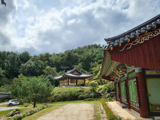 A traditional Korean building with ornate architectural details stands in a lush, green forested area. The roofs are intricately decorated, featuring vibrant colors and designs. A gravel path leads towards the main structure, and there's a white car parked nearby. The sky above is partly cloudy, adding a dynamic element to the serene setting.