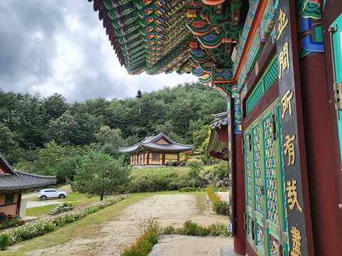 A traditional building with ornate, colorful wooden carvings is situated amidst lush greenery. The foreground features an intricately decorated wooden structure with visible Chinese characters. In the background, another building with a similar architectural style is surrounded by trees, and a parked white car is seen to the left.