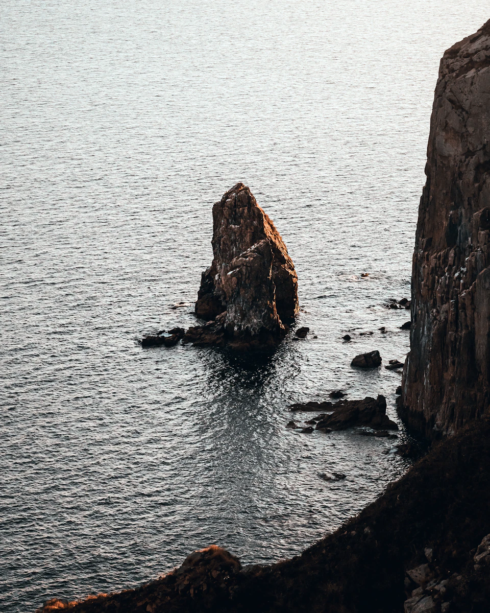 The image depicts a serene coastal scene with a large rock formation on the right side, its brown surface contrasting against the deep blue water surrounding it. The rock is situated at the edge of the ocean, where the water meets the land in a dramatic cliff face. In the foreground, there are several smaller rocks scattered across the water's surface, adding to the rugged beauty of the scene.