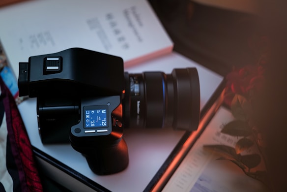 A digital camera with a large lens rests atop an open book. The camera's LCD screen displays settings, and the book's pages feature text and diagrams. Nearby, some flowers add a touch of color to the scene.