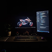 A dimly lit workspace featuring two computer monitors. One monitor displays an image of a motorcycle with striking red accents, while the other shows lines of computer code. A keyboard, mouse, and various desk items, including a laptop and a gaming controller, are arranged neatly. A plant adds a touch of greenery to the scene.