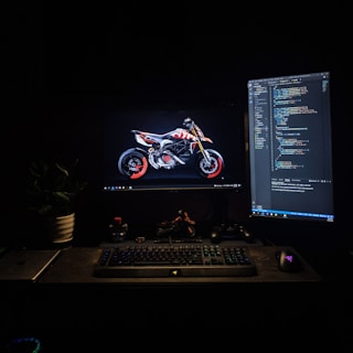 A dimly lit workspace featuring two computer monitors. One monitor displays an image of a motorcycle with striking red accents, while the other shows lines of computer code. A keyboard, mouse, and various desk items, including a laptop and a gaming controller, are arranged neatly. A plant adds a touch of greenery to the scene.