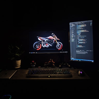 A dimly lit workspace featuring two computer monitors. One monitor displays an image of a motorcycle with striking red accents, while the other shows lines of computer code. A keyboard, mouse, and various desk items, including a laptop and a gaming controller, are arranged neatly. A plant adds a touch of greenery to the scene.