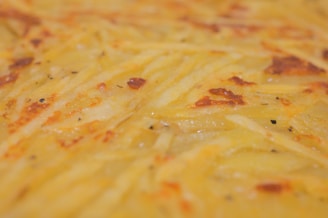 A close-up view of a golden-brown baked dish with a textured surface, likely made from thinly sliced potatoes with visible crusty edges. The surface seems to have a slightly greasy sheen, with hints of seasoning peppered throughout.