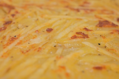 A close-up view of a golden-brown baked dish with a textured surface, likely made from thinly sliced potatoes with visible crusty edges. The surface seems to have a slightly greasy sheen, with hints of seasoning peppered throughout.