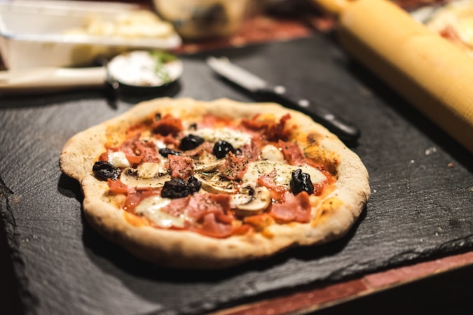 A freshly baked pizza with a golden-brown crust topped with slices of mushrooms, black olives, ham, and melted cheese. It rests on a dark slate serving board, surrounded by a knife, and a rolling pin suggesting a kitchen setting. The focus is on the pizza, with a slightly blurred background giving a warm and inviting ambiance.