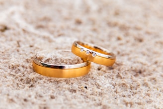 Two gold wedding rings are placed on a sandy surface. One of the rings is adorned with small diamonds, adding a touch of sparkle.