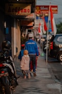 A man and two children walk down a sidewalk lined with shops and motorcycles. The man is wearing a blue 'CHELSEA' jacket and a white cap, while the children are dressed in colorful clothing. Several Indonesian flags are visible along the street, and various shop signs can be seen above.