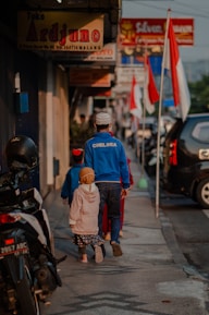 A man and two children walk down a sidewalk lined with shops and motorcycles. The man is wearing a blue 'CHELSEA' jacket and a white cap, while the children are dressed in colorful clothing. Several Indonesian flags are visible along the street, and various shop signs can be seen above.