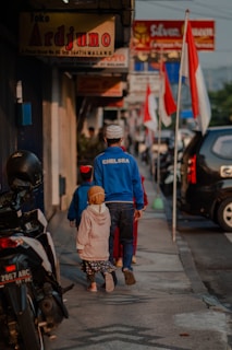 A man and two children walk down a sidewalk lined with shops and motorcycles. The man is wearing a blue 'CHELSEA' jacket and a white cap, while the children are dressed in colorful clothing. Several Indonesian flags are visible along the street, and various shop signs can be seen above.