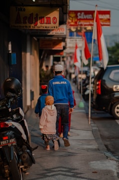 A man and two children walk down a sidewalk lined with shops and motorcycles. The man is wearing a blue 'CHELSEA' jacket and a white cap, while the children are dressed in colorful clothing. Several Indonesian flags are visible along the street, and various shop signs can be seen above.
