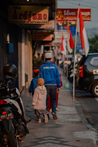 A man and two children walk down a sidewalk lined with shops and motorcycles. The man is wearing a blue 'CHELSEA' jacket and a white cap, while the children are dressed in colorful clothing. Several Indonesian flags are visible along the street, and various shop signs can be seen above.