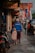A man and two children walk down a sidewalk lined with shops and motorcycles. The man is wearing a blue 'CHELSEA' jacket and a white cap, while the children are dressed in colorful clothing. Several Indonesian flags are visible along the street, and various shop signs can be seen above.