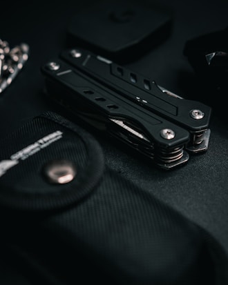 A multitool is surrounded by various dark-colored objects, including a textured black pouch. The scene is set against a dark backdrop, creating a sleek and minimalist aesthetic.