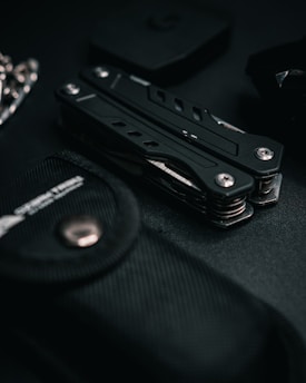A multitool is surrounded by various dark-colored objects, including a textured black pouch. The scene is set against a dark backdrop, creating a sleek and minimalist aesthetic.