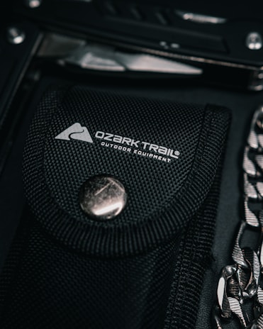 A black fabric sheath with a silver button closure and the Ozark Trail outdoor equipment logo is in focus. Nearby, partially visible, is a metallic tool and a chain.