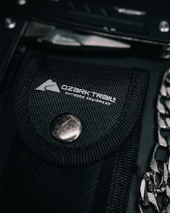 A black fabric sheath with a silver button closure and the Ozark Trail outdoor equipment logo is in focus. Nearby, partially visible, is a metallic tool and a chain.