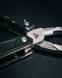 A close-up of a multi-tool with a focus on the plier component. The tool features dark handles and a metallic finish with visible screws and a small icon engraved on the metal surface. The background is a dark, textured material.