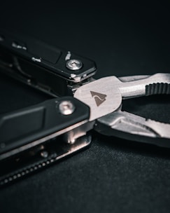 A close-up of a multi-tool with a focus on the plier component. The tool features dark handles and a metallic finish with visible screws and a small icon engraved on the metal surface. The background is a dark, textured material.