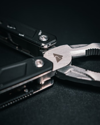 A close-up of a multi-tool with a focus on the plier component. The tool features dark handles and a metallic finish with visible screws and a small icon engraved on the metal surface. The background is a dark, textured material.