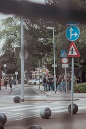 A group of people gather near a street corner with various traffic signs. The scene includes lush green trees, a pedestrian crossing, and a parked white car in the background. The atmosphere is casual and lively, with individuals appearing to chat and interact.