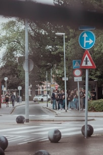 A group of people gather near a street corner with various traffic signs. The scene includes lush green trees, a pedestrian crossing, and a parked white car in the background. The atmosphere is casual and lively, with individuals appearing to chat and interact.
