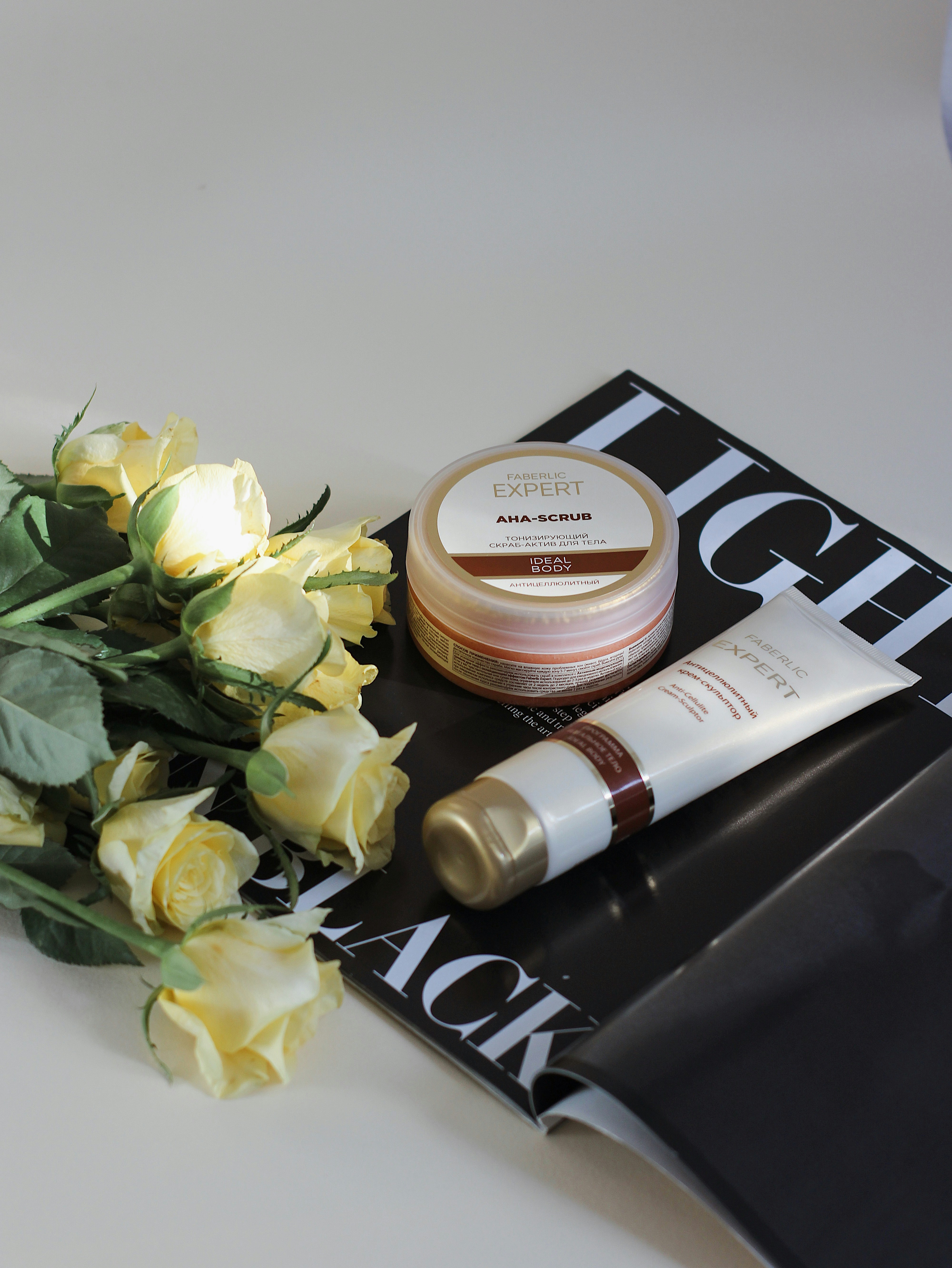 A set of skincare products including a jar of AHA scrub and a tube of cream. They are placed on top of an open black and white magazine. Beside the products, there is a bouquet of light yellow roses. The scene is set against a light background, with soft lighting highlighting the items.