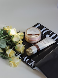 A set of skincare products including a jar of AHA scrub and a tube of cream. They are placed on top of an open black and white magazine. Beside the products, there is a bouquet of light yellow roses. The scene is set against a light background, with soft lighting highlighting the items.