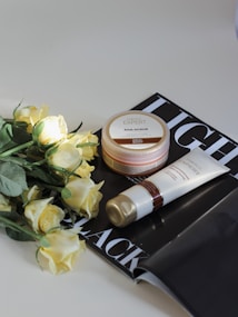A set of skincare products including a jar of AHA scrub and a tube of cream. They are placed on top of an open black and white magazine. Beside the products, there is a bouquet of light yellow roses. The scene is set against a light background, with soft lighting highlighting the items.