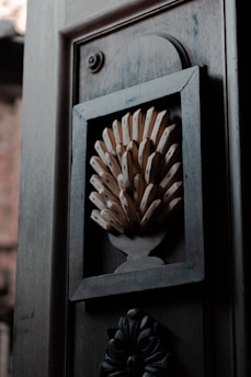 A wooden door panel features an intricate decorative element, resembling a stylized, abstract bouquet or fan of wooden objects enclosed in a rectangular frame. The dark wood and shadows give the design a textured and three-dimensional appearance.