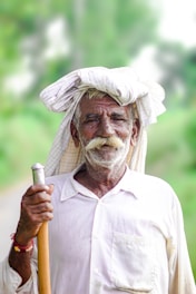An elderly man with a white mustache wears a white shirt and a traditional head wrap while holding a wooden stick. He stands outdoors against a blurred green background, exuding a sense of wisdom and calmness.