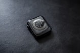 A smartwatch with a circular sensor design placed face down on a textured dark surface. The watch has a sleek, rounded design with a metallic rim and visible screws surrounding the sensor area.