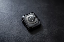A smartwatch with a circular sensor design placed face down on a textured dark surface. The watch has a sleek, rounded design with a metallic rim and visible screws surrounding the sensor area.