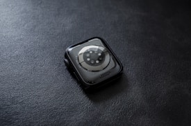 A smartwatch with a circular sensor design placed face down on a textured dark surface. The watch has a sleek, rounded design with a metallic rim and visible screws surrounding the sensor area.