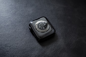 A smartwatch with a circular sensor design placed face down on a textured dark surface. The watch has a sleek, rounded design with a metallic rim and visible screws surrounding the sensor area.