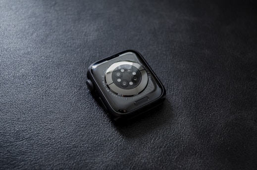 A smartwatch with a circular sensor design placed face down on a textured dark surface. The watch has a sleek, rounded design with a metallic rim and visible screws surrounding the sensor area.