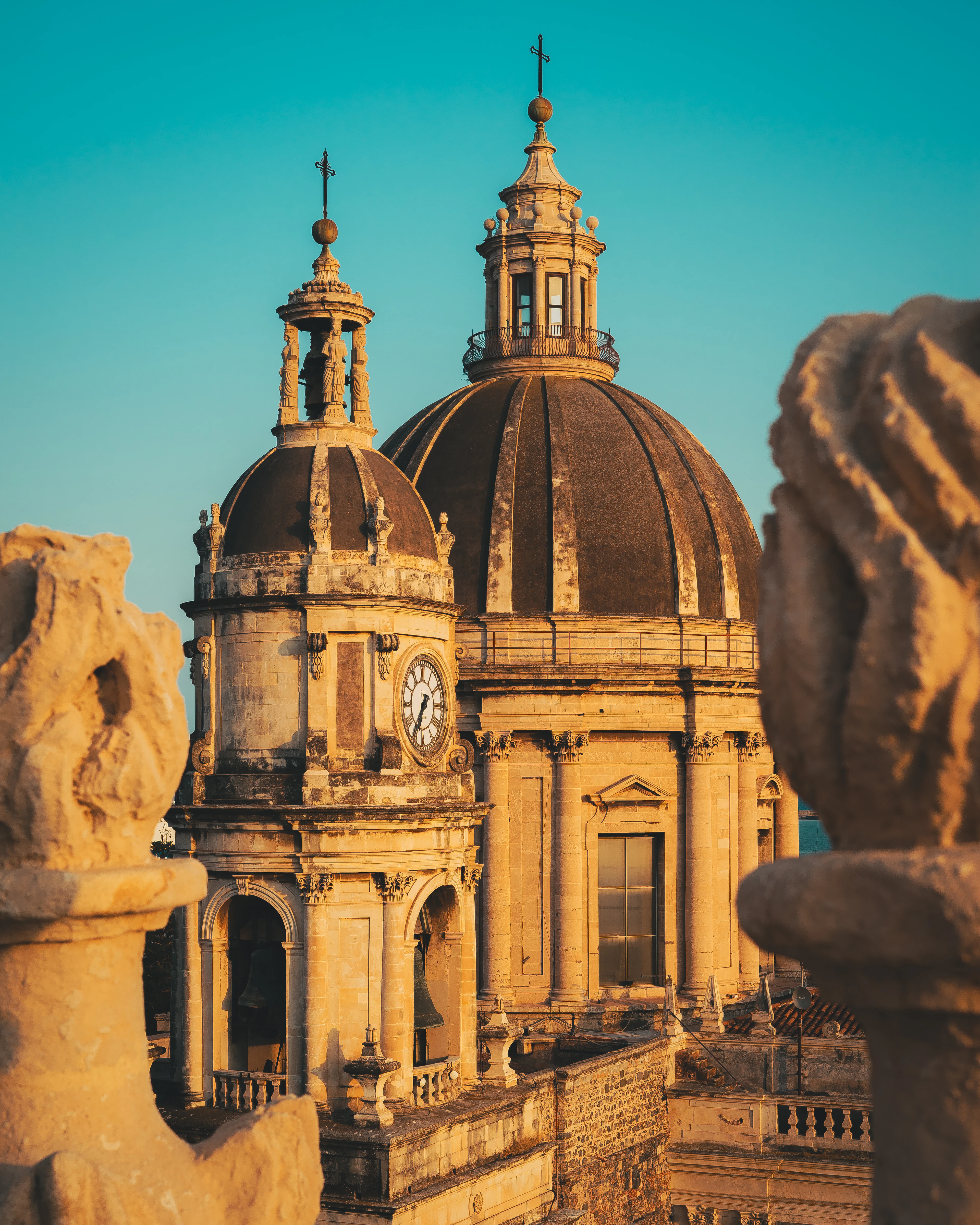 Weathered baroque domes and a clock tower rising above Catania's rooftops, framed by carved stone balustrades in warm evening light