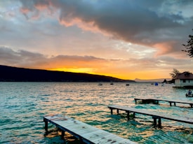 A serene lakeside view during sunset, with a vibrant sky colored in shades of orange and pink reflecting on the water. The silhouette of distant hills creates a striking contrast against the warm colors. Wooden docks extend into the lake, and small boats are visible floating near the horizon. A house with trees is situated on the right side, adding a peaceful ambiance.