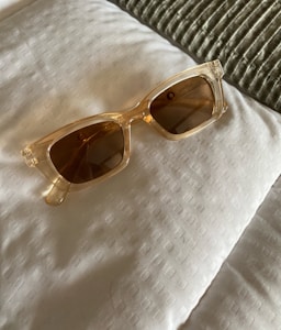 A pair of transparent, square-framed sunglasses with brown lenses, placed on a white, textured fabric surface. The fabric appears to be part of a bed, judging by the visible pillow and the greenish-gray, ribbed textile in the background.