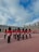 A group of uniformed guards in bright red coats and characteristic tall black hats stands in formation in a large open courtyard. The backdrop includes an old stone building with arched doorways and decorative chimneys under a partly cloudy sky.