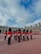 A group of uniformed guards in bright red coats and characteristic tall black hats stands in formation in a large open courtyard. The backdrop includes an old stone building with arched doorways and decorative chimneys under a partly cloudy sky.