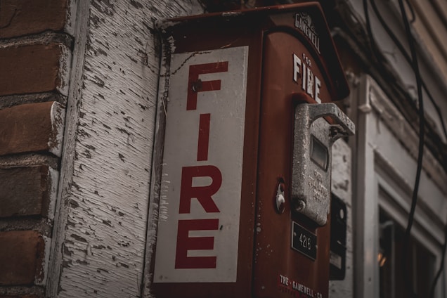 A vintage fire alarm box mounted on a wall, featuring a rusted red exterior with the word 'FIRE' in bold white letters. The surrounding area shows peeling white paint and a brick surface, giving the scene an aged, urban feel.