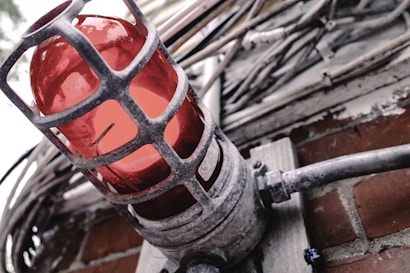 A red industrial warning light encased in a metal protective cage is mounted on a brick wall. Surrounding the light are several thick cables and conduits, suggesting an industrial or mechanical setting. The metal of the cage is weathered, adding a rugged character to the scene.
