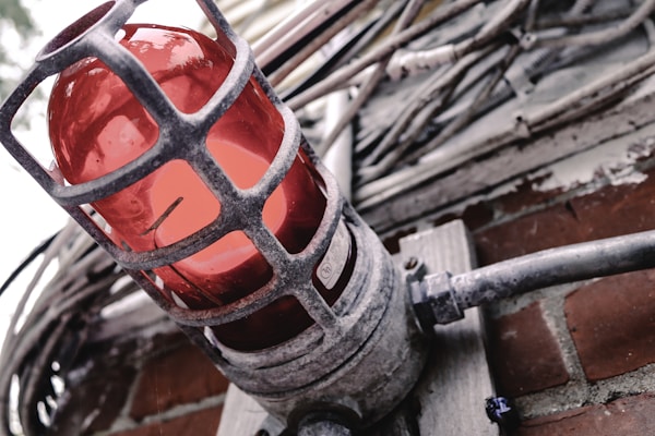 A red industrial warning light encased in a metal protective cage is mounted on a brick wall. Surrounding the light are several thick cables and conduits, suggesting an industrial or mechanical setting. The metal of the cage is weathered, adding a rugged character to the scene.