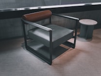A modern armchair with a sleek black frame, featuring a brown cylindrical leather headrest and a dark gray cushioned seat. Next to the chair is a small round side table with a textured white surface and green geometric legs. The setting has a minimalist and contemporary design with a neutral-toned carpeted floor and a concrete wall in the background.